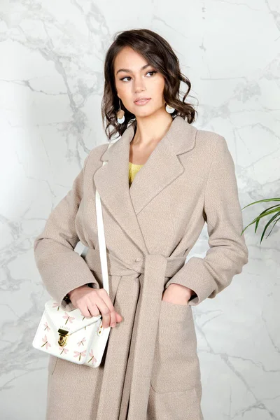 Beauty Fashion Model Girl with curls in light beige coat in a bright white loft style studio. Beautiful Luxury Winter Woman. Beauty and fashion. Spring and autumn clothes fashion Girl posing at studio