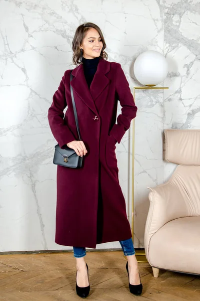 Beauty Fashion Model Girl with curls in burgundy coat in a bright white loft style studio. Beautiful Luxury Winter Woman. Beauty and fashion. Spring and autumn clothes fashion. Girl posing at studio.