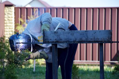Kaynak maskeli, gri kazaklı, fermuarlı, lacivert pantolonlu ve eldivenli bir adam. Kaynak makinesi açık havada, kırmızı kahverengi metal, eğri büğrü tahta çitlerin ve mavi gökyüzünün önünde.