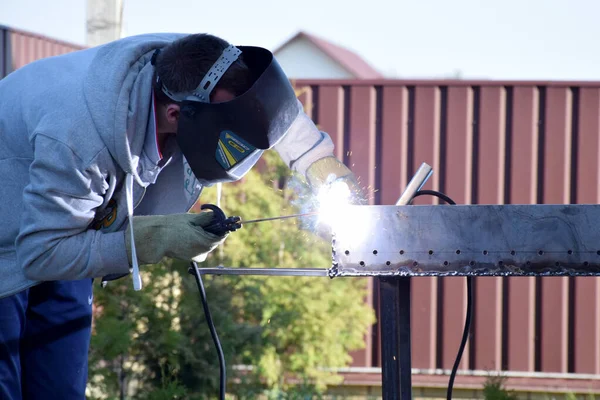 Man in protecting welding helmet mask grey sweater with zipper dark blue pants and gauntlet work with welding machine outdoors in front of red brown metal corrugated board fence and blue sky