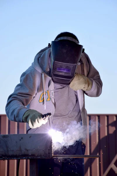 Man in protecting welding helmet mask grey sweater with zipper dark blue pants and gauntlet work with welding machine outdoors in front of red brown metal corrugated board fence and blue sky