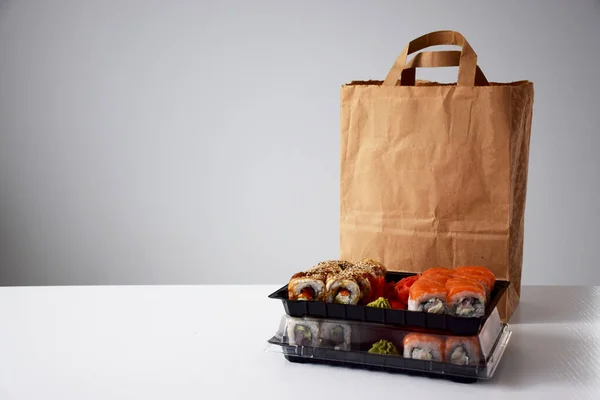 Suşi beyaz masadaki kağıt paketin yanındaki plastik kutuda yuvarlanıyor. Teslimat. Götürün.
