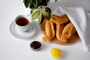 Beyaz mutfak peçetesinin altında taze çıtır çörekler ahşap kesim tahtası, reçel, bal, beyaz masada kahvaltıda servis edilen sıcak çay.