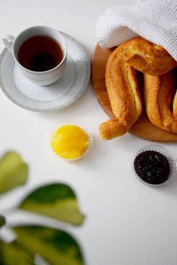 Tahta kesme tahtasındaki beyaz mutfak havlusunun altında taze gevrek rulo tabakta siyah çay, kahvaltıda reçel ve bal.