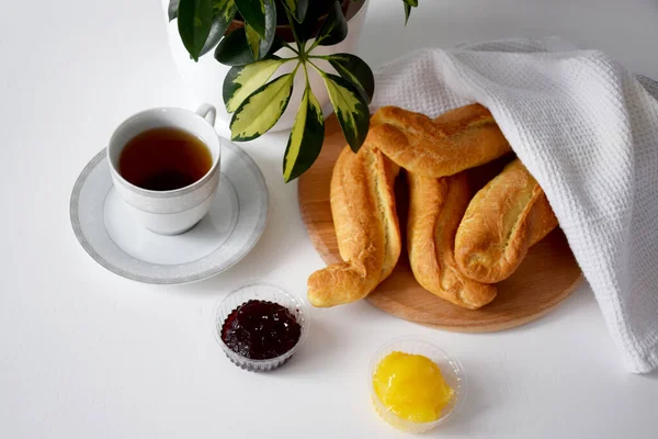 Beyaz mutfak peçetesinin altında taze çıtır çörekler ahşap kesim tahtası, reçel, bal, beyaz masada kahvaltıda servis edilen sıcak çay.