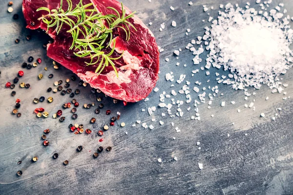 Beef steak with spilled colored pepper, sea salt and sprig of rosemary on the gray structured background