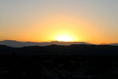 Bir çöl dağı manzarası, parlak sarı ve turuncu gün batımı siluetinde.