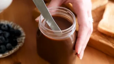 Çikolatalı fındık ezmesi ya da kavanozda tereyağı. Bir parça çikolatalı fındık ezmesini ekmek ekmeğine sürmek için masa bıçağıyla alan kişi.