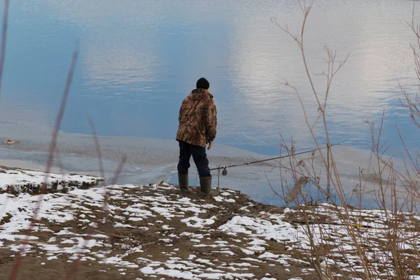 一个穿着迷彩服的渔夫站在河岸上 手里拿着一根钓竿 天气很冷 地上可以看到雪 — 图库照片