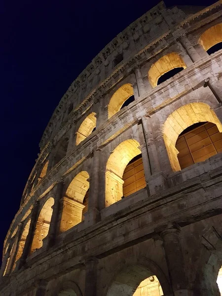 Close-up of Colosseum wall with lights on against dark blue sky.