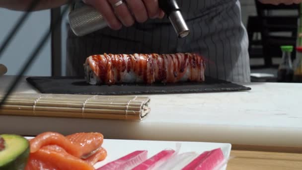 新鮮なおいしい寿司ロールを用意する寿司マスターは サーモンをカラメル化するためにトーチを使用しています コンセプト 竹の間に巻き寿司ロールを転がす — ストック動画