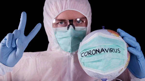 Man in professional white safety uniform is showing peace sign, holding infected globe with coronavirus text on green mask. Covid-19 pandemic. World epidemic.
