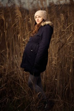 Ormanda hamile bir kadın ya da sonbaharda ya da kışın çimen. Çimlerin üzerinde duran bir kadının hamilelik fotoğrafı. Şapka ceketini soğuk giyer.