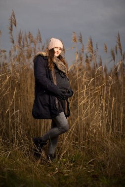 Ormanda hamile bir kadın ya da sonbaharda ya da kışın çimen. Çimlerin üzerinde duran bir kadının hamilelik fotoğrafı. Şapka ceketini soğuk giyer.