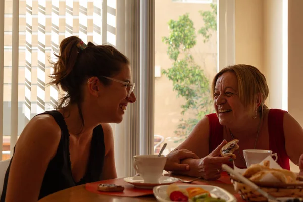 two persons adult and young beautiful woman laughing drinking coffee and eating healthy food at home in the afternoon