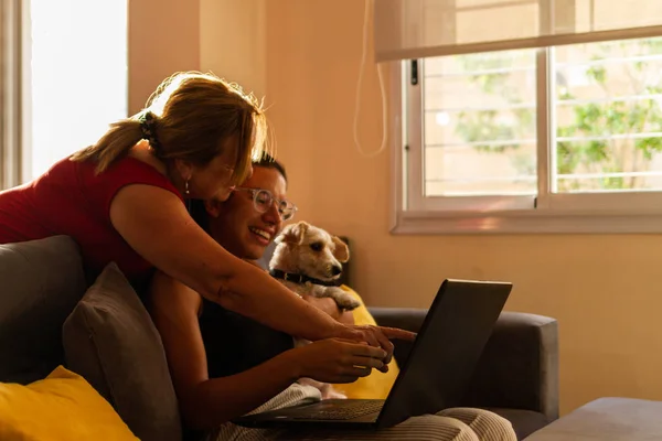 adult and young latin and beautiful woman chatting happily at home using laptop sitting at grey couch with dog