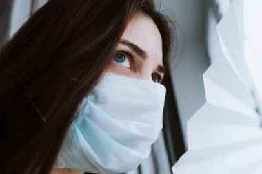 Tıbbi koruyucu maskeli genç bir kadın pencereden dışarı bakıyor..
