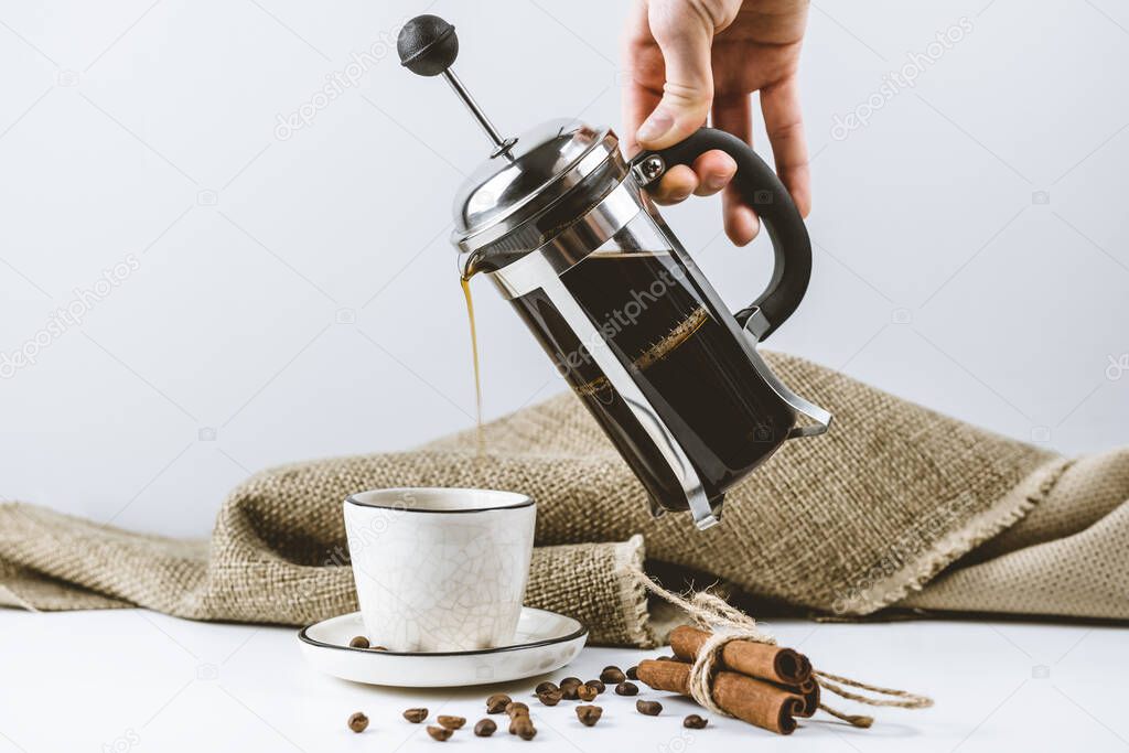 A female hand pours brewed coffee in a French press into a white handmade cup. 
