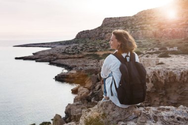 Gezgin güzel stil sahibi kadın deniz manzaralı yüksek kayalarda oturur. Şafağı ya da gün batımını bekliyor.
