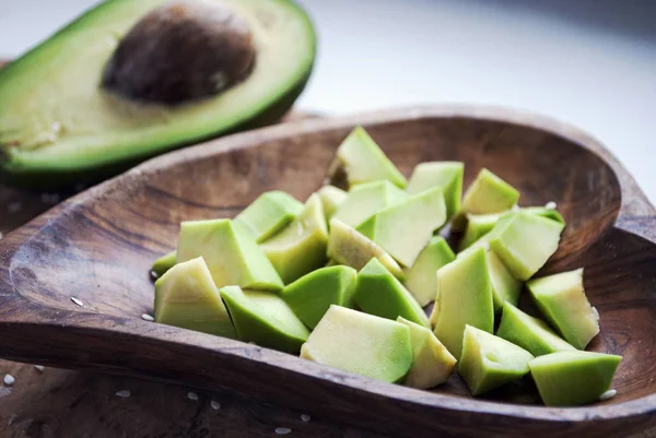 Chopped avocado are in a wooden dish in the form of a heart next to half avocado.