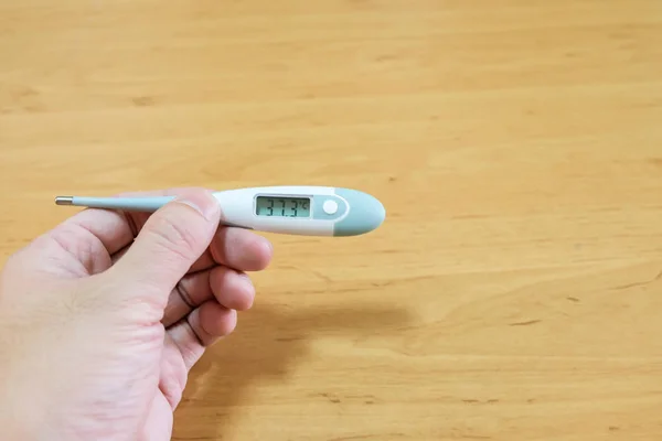 Men hand holding digital thermometer to measure body temperature and check for fever on wood background. Sickness, healthcare and medical concept.
