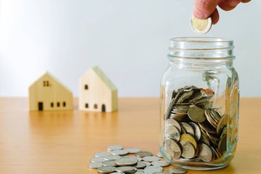 Erkeklerin seçici odak noktası, ev için biriktirmek için cam kavanoza para koymak, arka planda küçük ahşap ev olan bir mülk. Ev kredisi için servet ve tasarruf planı. Kişisel yatırım kavramı.