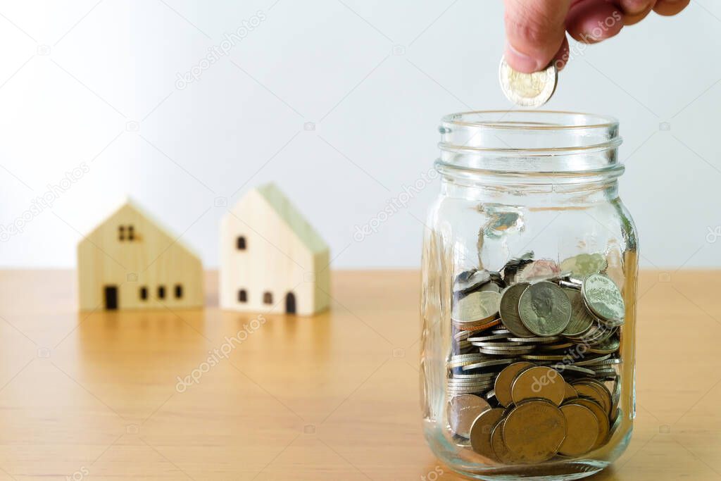 Selective focus of Men hand puting coin into glass jar for saving for home, property with small wood house on the background. Wealth and saving plan for house mortgage. Personal investment concept.