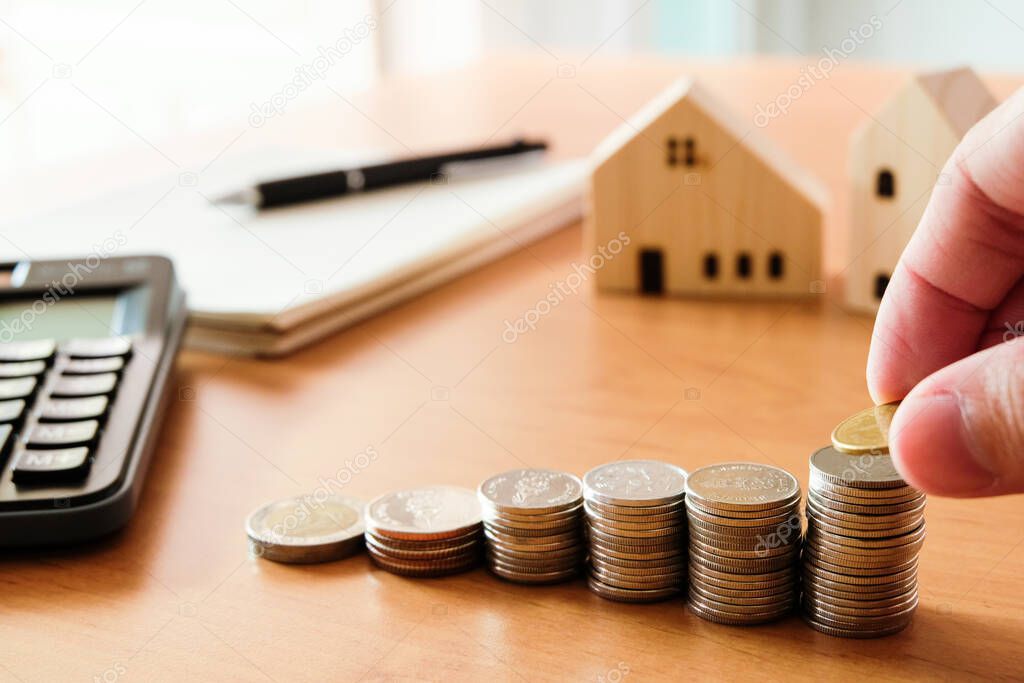 Selective focus Men hand puting coin on stack of money coin for saving for home, property with small wood house on the background. Wealth and saving plan for house mortgage.Personal investment concept
