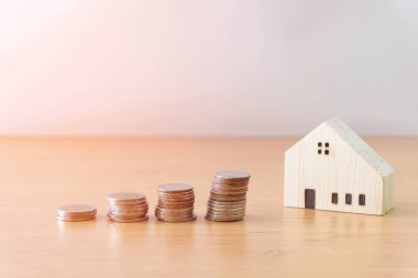 Para biriktirme yığını ahşap masadaki küçük ahşap eve ulaşmak için artıyor. Arka planı beyaz ve metin için yeri var. Ev ya da mülk için kişisel finansman. Bütçe planlama.