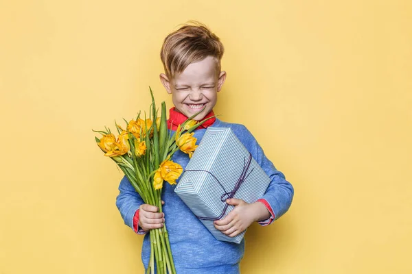 Menino bonito segurando cacho de flores e caixa de presente. Dia dos Namorados. Aniversário. Dia da mãe. Retrato de estúdio sobre fundo amarelo — Fotografia de Stock