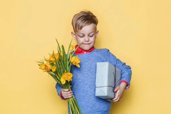 Menino bonito segurando cacho de flores e caixa de presente. Dia dos Namorados. Aniversário. Dia da mãe. Retrato de estúdio sobre fundo amarelo — Fotografia de Stock