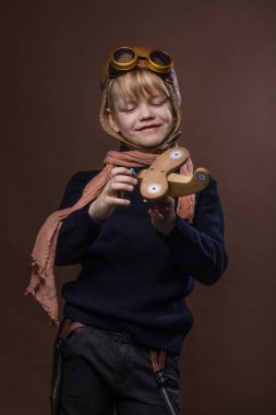 Mutlu çocuk pilot şapka ve gözlük giymiş. Ahşap oyuncak uçak ile oynayan çocuk. Rüya ve özgürlük kavramı. Retro tonda. Stüdyo portre kahverengi zemin üzerinde