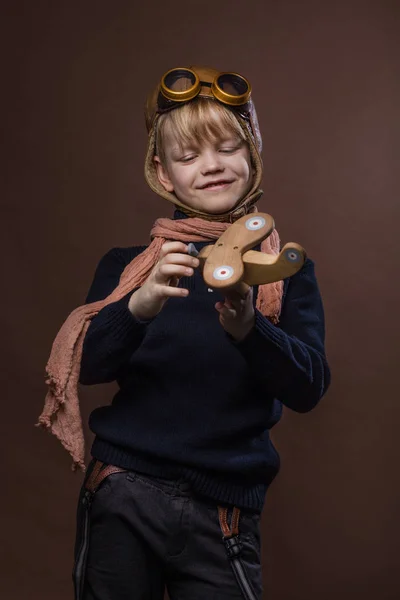 Niño feliz vestido con sombrero piloto y gafas. Niño jugando con el avión de juguete de madera. Concepto sueño y libertad. Retro tonificado. Estudio retrato sobre fondo marrón — Foto de Stock