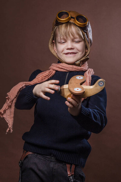 Счастливый ребенок в пилотской шляпе и очках. Ребёнок играет с деревянным игрушечным самолётом. Концепция мечты и свободы. Ретро тонизирован. Студийный портрет на коричневом фоне
