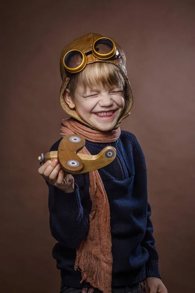 Criança feliz vestida de chapéu piloto e óculos. Criança brincando com avião de brinquedo de madeira. Conceito de sonho e liberdade. Retro tonificado. Retrato de estúdio sobre fundo marrom — Fotografia de Stock