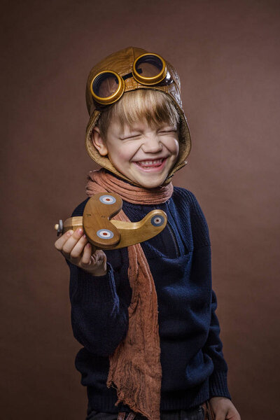 Happy child dressed in pilot hat and glasses. Kid playing with wooden toy airplane. Dream and freedom concept. Retro toned. Studio portrait over brown background