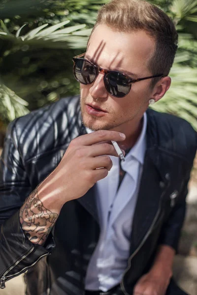 Jeune homme à la mode avec des lunettes de soleil et une veste en cuir fumant la cigarette. Portrait extérieur — Photo