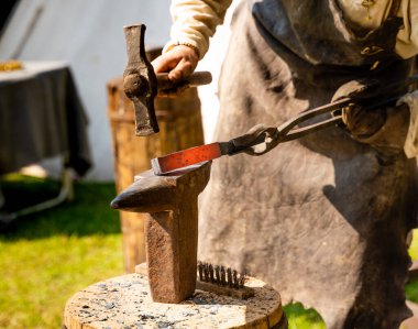 Bir demirci açık hava ocağında kene ve demir ocakları olan bir eser tutar. Eller yukarı. Yaşayan Tarih Festivali 'nde Ortaçağ Demircilik Atölyesi