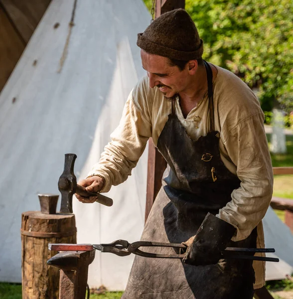 A blacksmith  forges in an open-air forge. Smiling face of pretty worker. Middle Ages Blacksmithing Workshop at Living History Festival