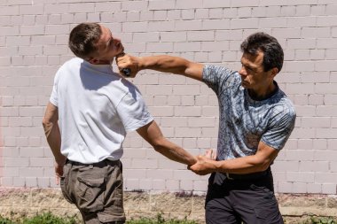Dövüş sanatları eğitmenleri Krav Maga sokak dövüşlerinde kendini savunma tekniklerini gösterirler. Savunma, bir saldırganın yüzüne elinden alınan bir silahla vurur.
