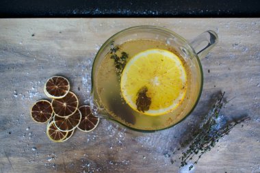 Sarı meyve çayı, bir dilim limon ve lezzet ve biberiye, cam bir kasede, üst manzara. Kurumuş narenciye meyveleri arka plandaki ahşap bir masanın üzerine serpilmiş toz serpiştirilmiş.