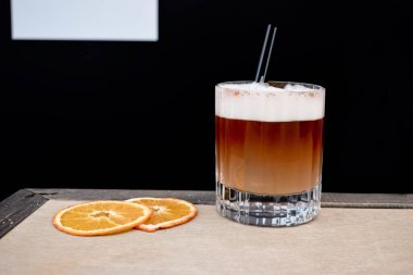 Köpükle kaplı kahverengi bir kokteyl siyah arka plandaki ahşap bir masada duruyor. Yakınlarda kuru bir portakal yatıyor.