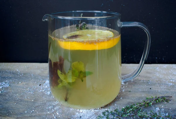 Tarçınlı sarı limon çayı ve siyah duvara karşı cam çaydanlıkta nane şekeri. Zitrus ve sopalar ağlar, bir içkinin içinde yüzer. Tahta bir masanın üzerinde bir biberiye filizi