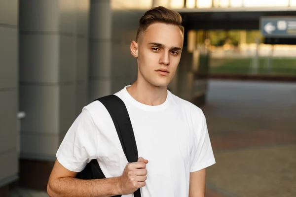 Retrato de un joven guapo de moda en una camisa blanca con una mochila cerca del edificio moderno . — Foto de Stock