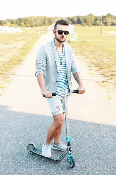 Elegante bell'uomo in abiti alla moda in sella a uno scooter Kick sulla spiaggia — Foto Stock