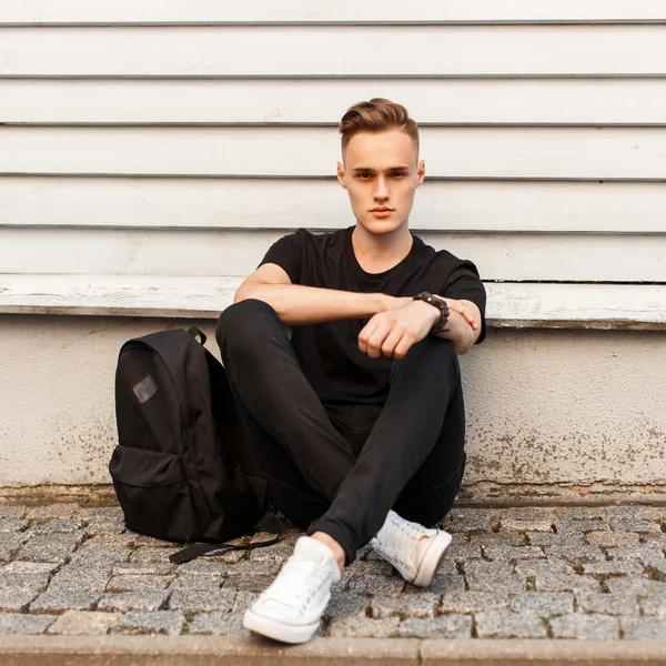 Joven hombre con estilo en camisa negra y zapatos blancos con una mochila se sienta cerca de la pared de madera — Foto de Stock