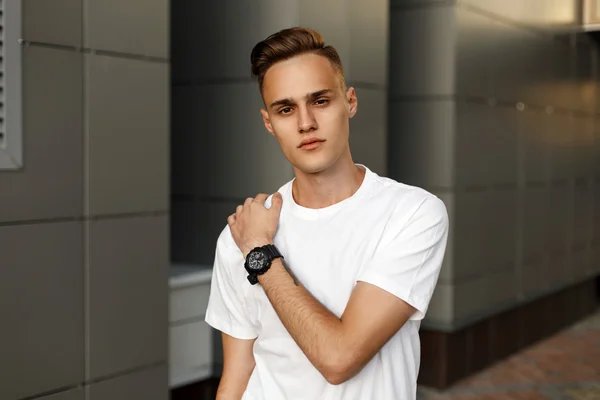 Joven chico guapo en una camiseta blanca posando cerca de la pared gris moderna . — Foto de Stock