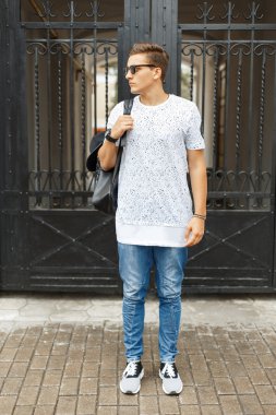 Beyaz tişörtlü, kot pantolonlu ve spor ayakkabılı genç ve yakışıklı bir hippi sokakta dikiliyor. Omzunda şapka olan bir sırt çantası..