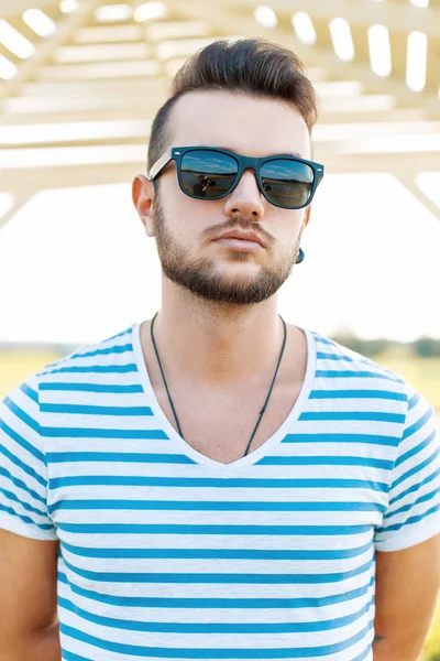 Knappe man in een t-shirt met de baard in zonnebril staande op het strand. — Stockfoto