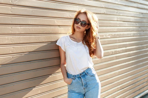 Стильная красивая хипстерша в солнцезащитных очках и винтажных джинсах, прислонившихся к деревянной стене — стоковое фото
